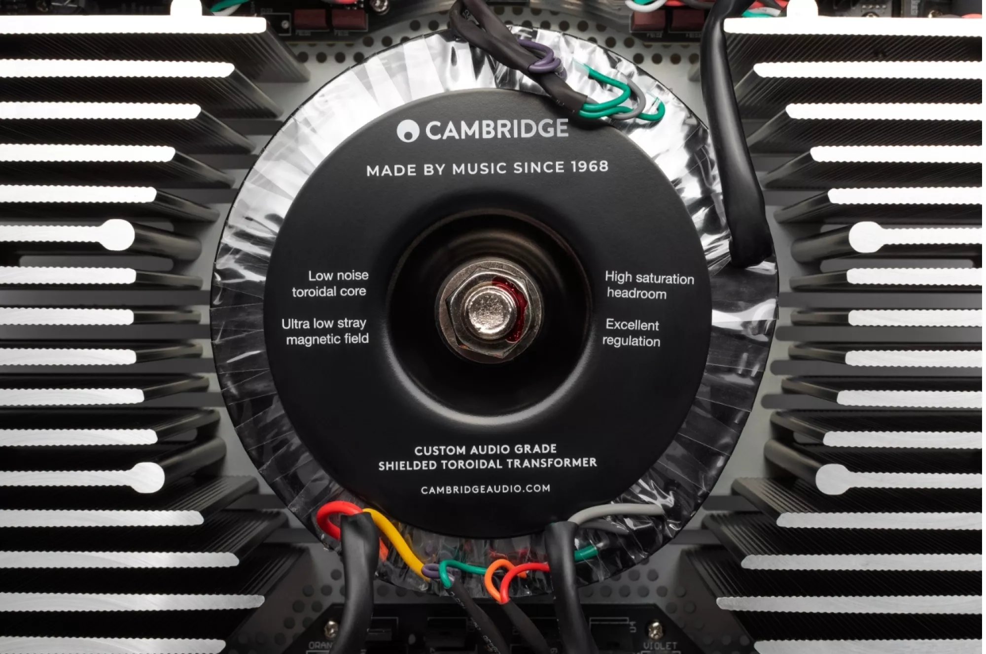 Detailní pohled na černý stíněný toroidní transformátor Cambridge s nápisem Made by Music since 1968, obklopený hliníkovými chladiči