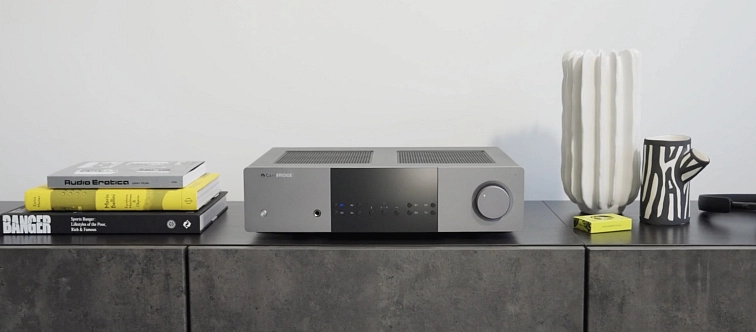 Cambridge Audio EXA100 v šedém provedení na tmavém betonovém komodě, vedle stohu knih a designové vázy