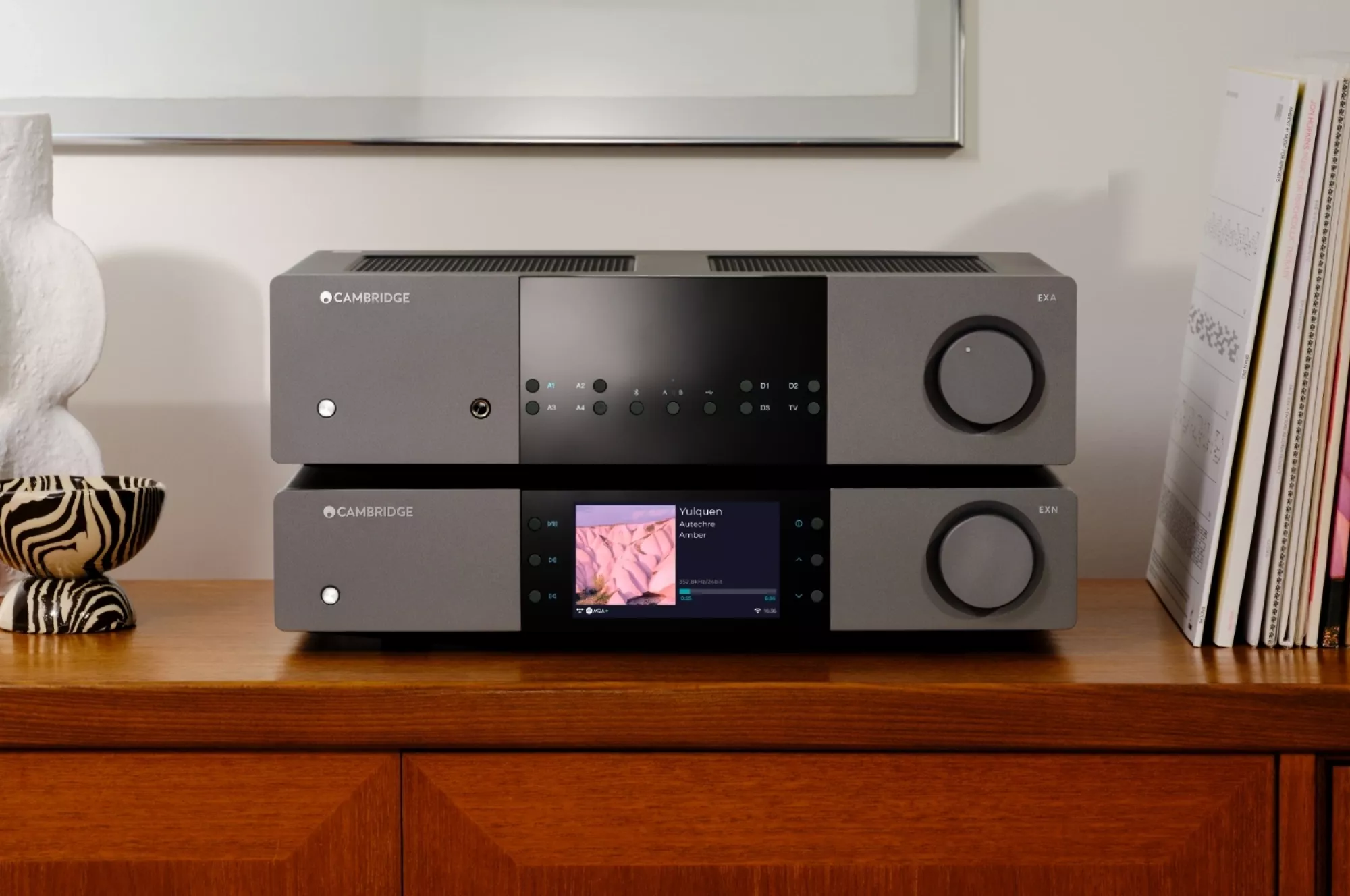 Cambridge Audio EXA100 postavený na síťovém přehrávači EXN100 na teakové komodě, na displeji streamera běží hudba