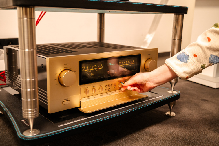 Detail integrovaného zesilovače Accuphase E-4000 — zlatá čelní deska s ručičkovými VU metry, ruka vysouvá spodní výklopný panel s ovládacími tlačítky