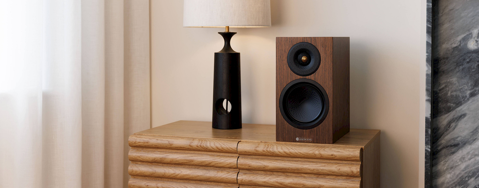 Monitor Audio Silver 50 8G v dýze European Walnut na dřevěné komodě vedle černé stolní lampy
