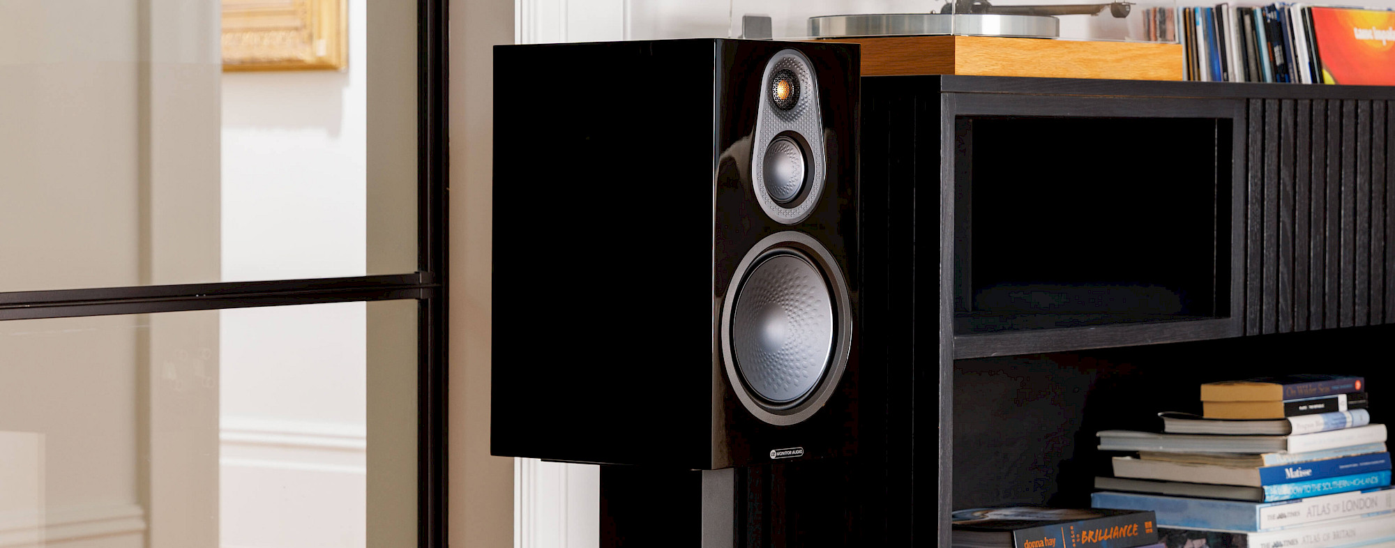 Monitor Audio Silver 100 8G v provedení High Gloss Black na stojanu vedle nábytkové sestavy s gramofonem a knihami