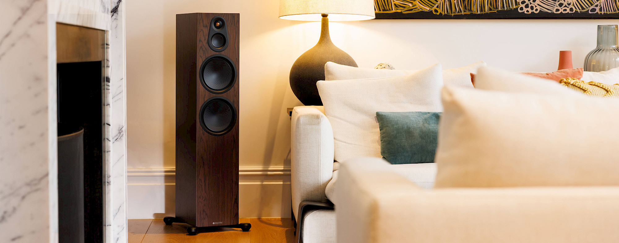 Sloupová reprosoustava Monitor Audio Silver 500 8G v provedení European Walnut vedle světlé pohovky, stojací lampy a mramorového krbu