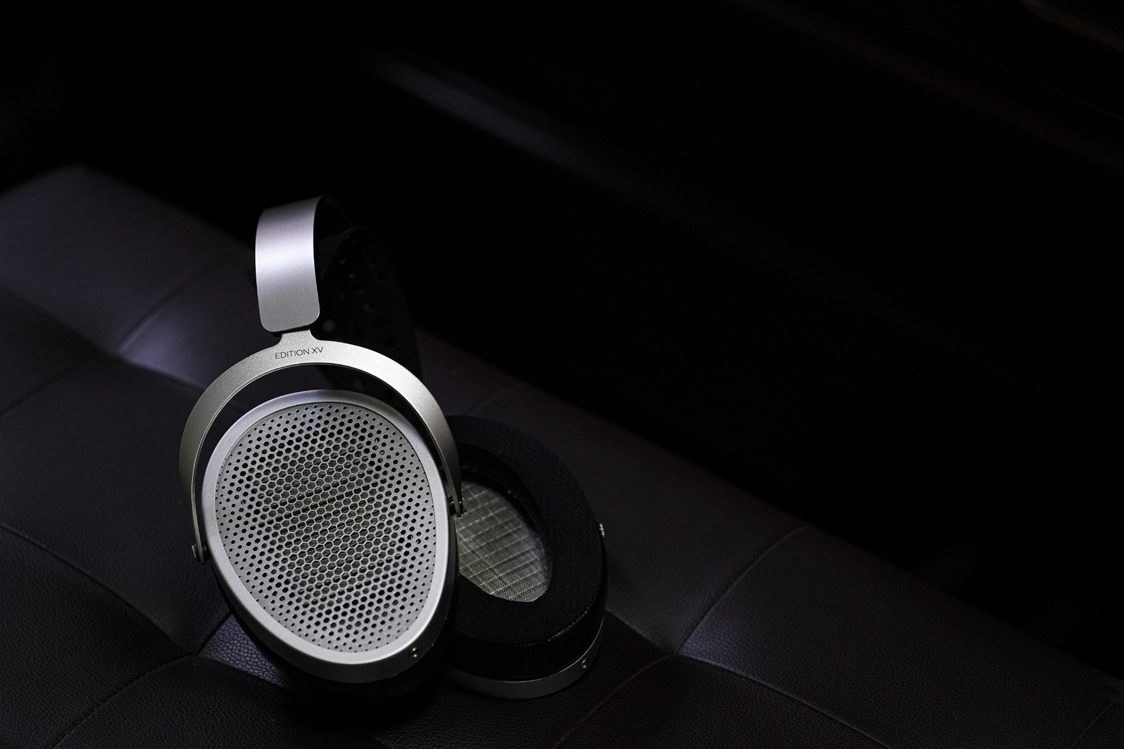 Sluchátka HIFIMAN Edition XV položená na tmavé kožené sedačce, detail stříbrné mřížky a náušníku