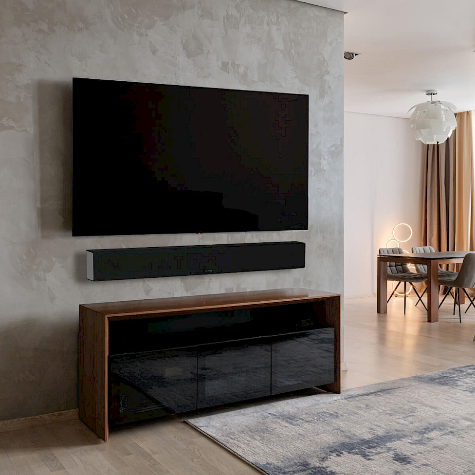 Soundbar Monitor Audio pod televizorem na betonové stěně nad komodou z ořechu