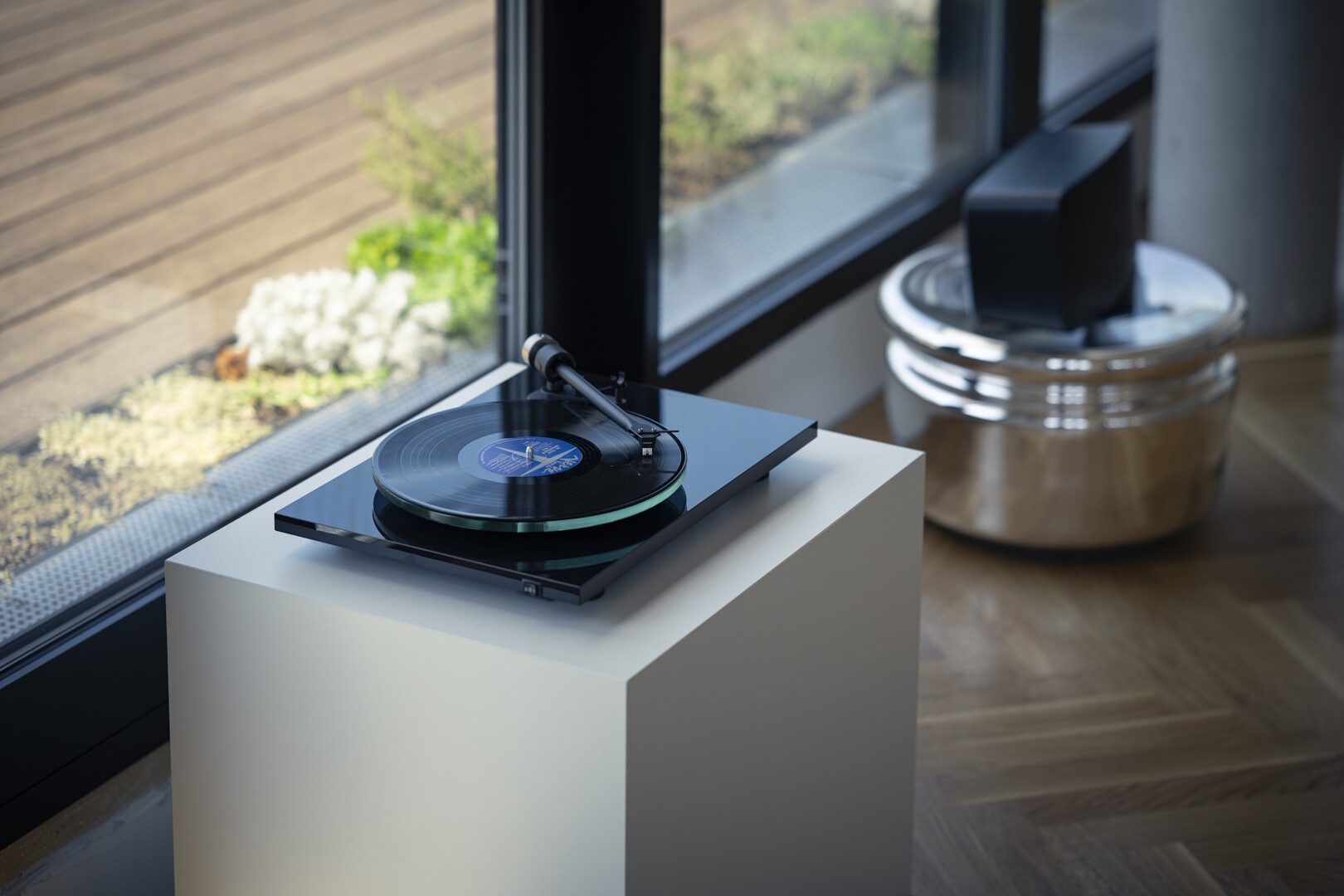 Gramofon Pro-Ject T2 v provedení piano černá s hrající deskou na bílém soklu u prosklené stěny obývacího pokoje