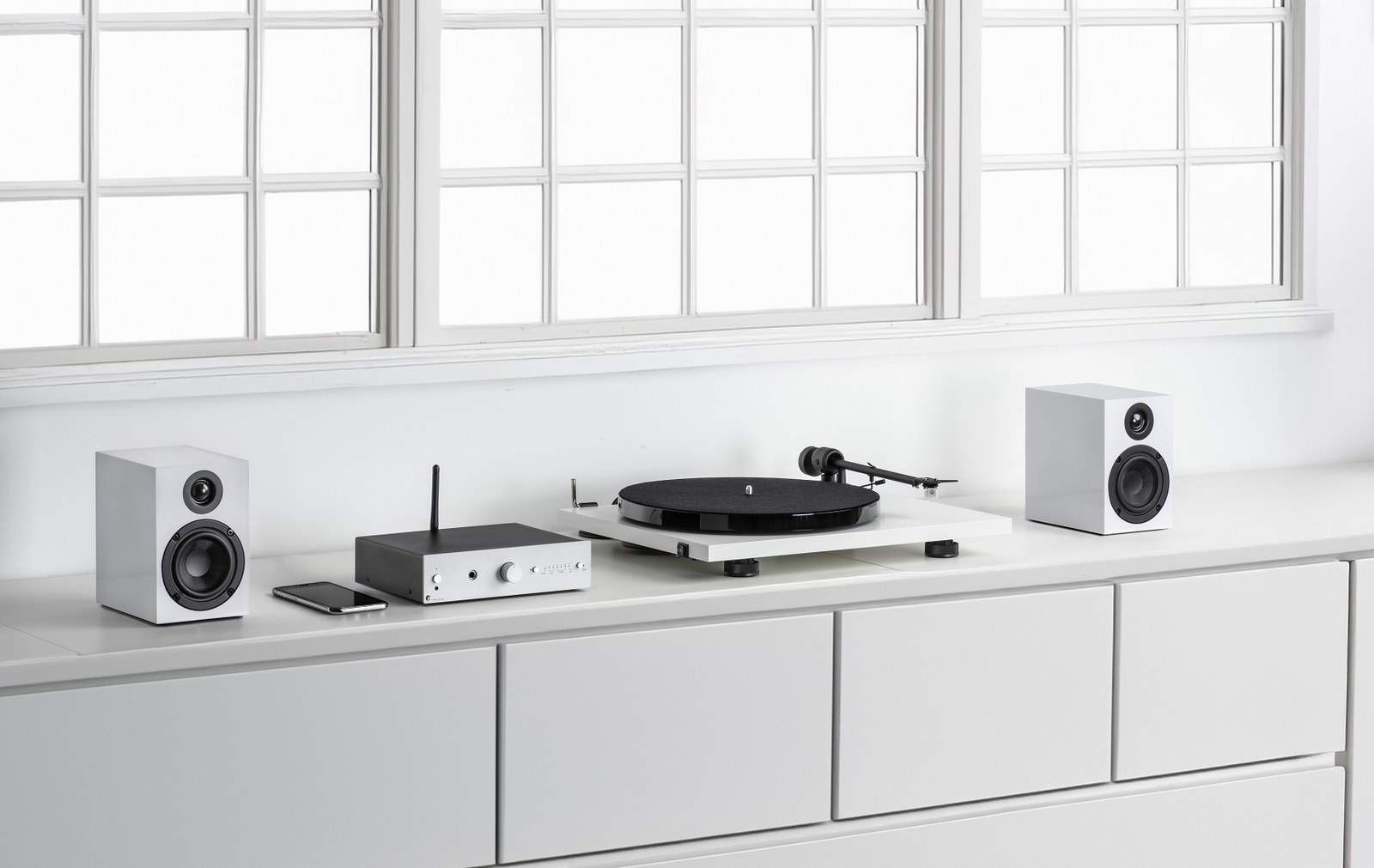Bílé provedení sestavy Pro-Ject Stereo Set E Phono na bílé komodě pod okny, gramofon uprostřed a obě reprosoustavy po stranách