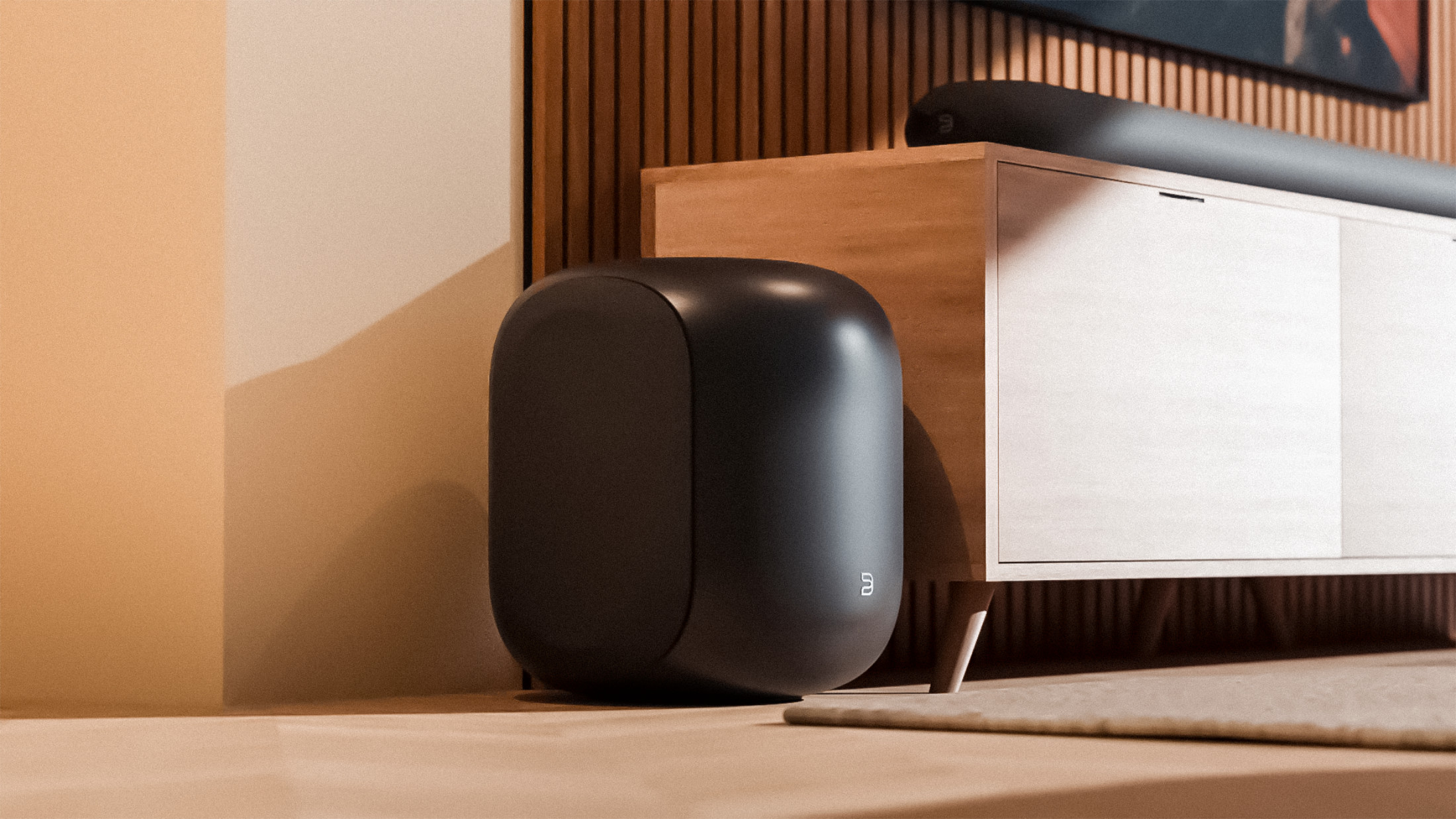 Subwoofer Bluesound PULSE SUB stojí vedle světlé dřevěné komody v teplém večerním světle