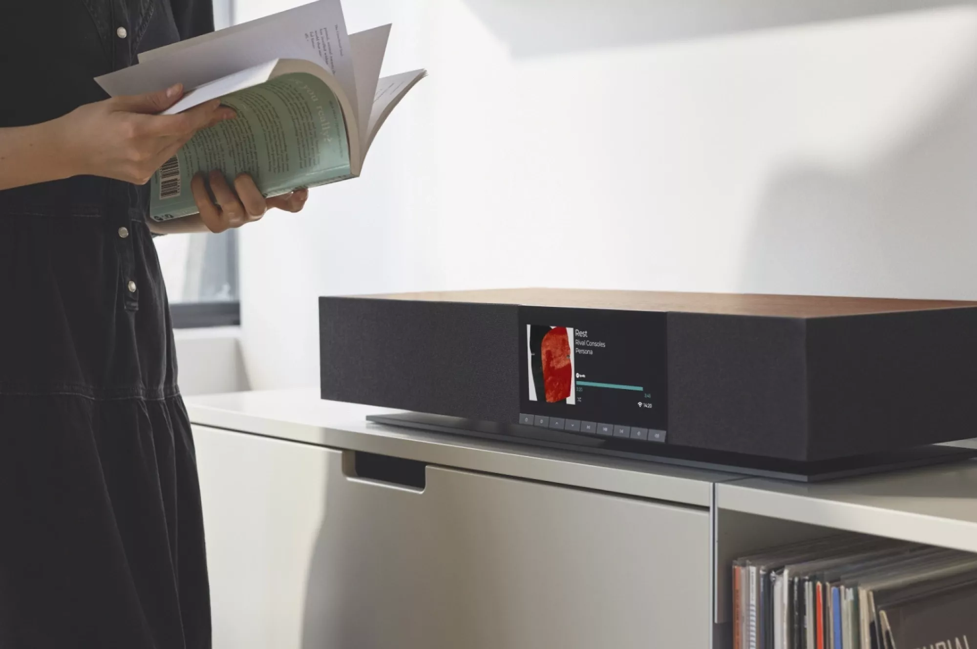 Detail čelního displeje Cambridge Audio Evo One s informacemi o přehrávané skladbě, vedle stojící žena listuje knihou a v komodě jsou vinylové desky