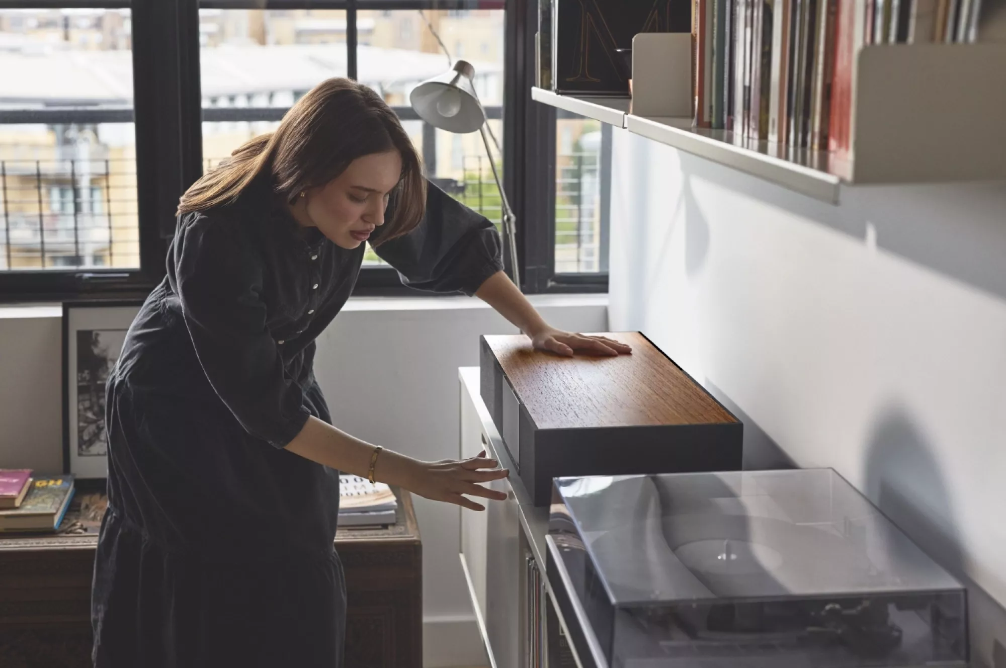 Žena v tmavých šatech pokládá ruku na dřevěný vrch systému Cambridge Audio Evo One umístěného na bílé komodě, vedle gramofon a regál s vinylovými deskami u okna