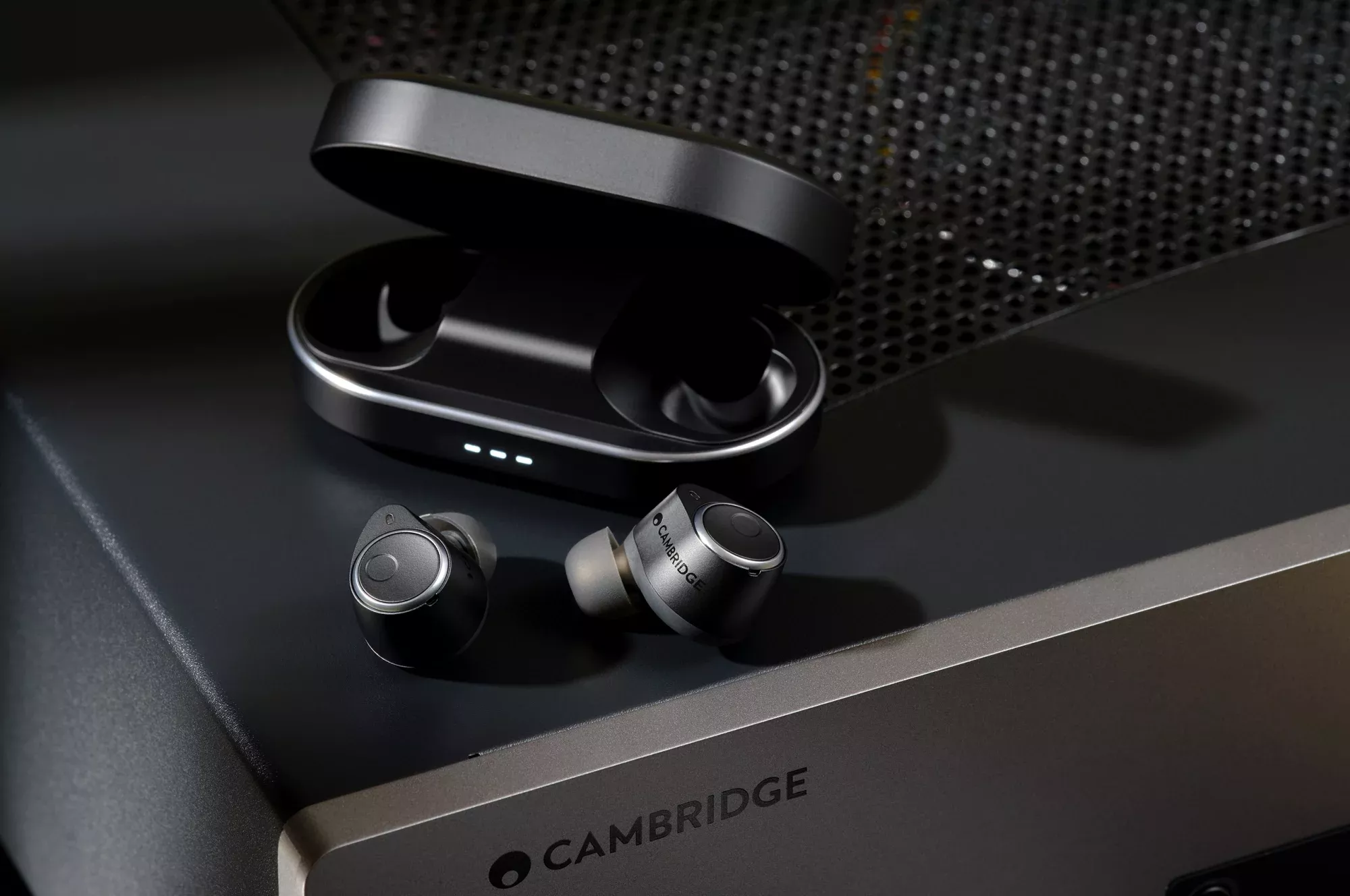 Sluchátka Cambridge Audio Melomania M100 a otevřené nabíjecí pouzdro položené na stolním zesilovači Cambridge