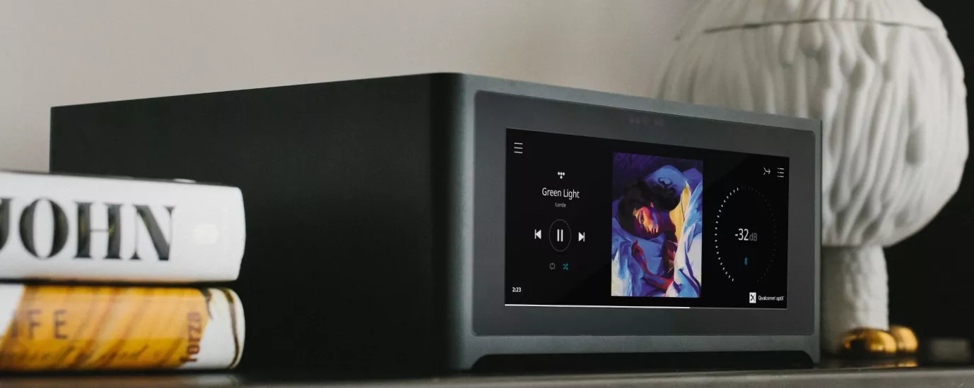 NAD M10 V3 postavený na konzolovém stolku vedle stohu knih a stolní lampy, na displeji běží obal alba