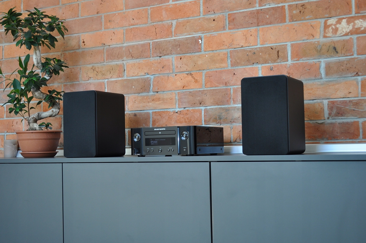 Marantz Melody X mezi dvojicí černých reprosoustav na šedé komodě před cihlovou stěnou