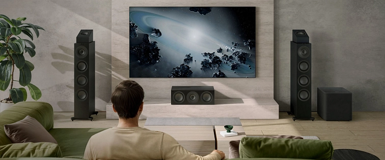 Domácí kino v obývacím pokoji, muž na zelené pohovce sleduje vesmírnou scénu na velké televizi, po stranách sloupové reproduktory, středový kanál a subwoofer