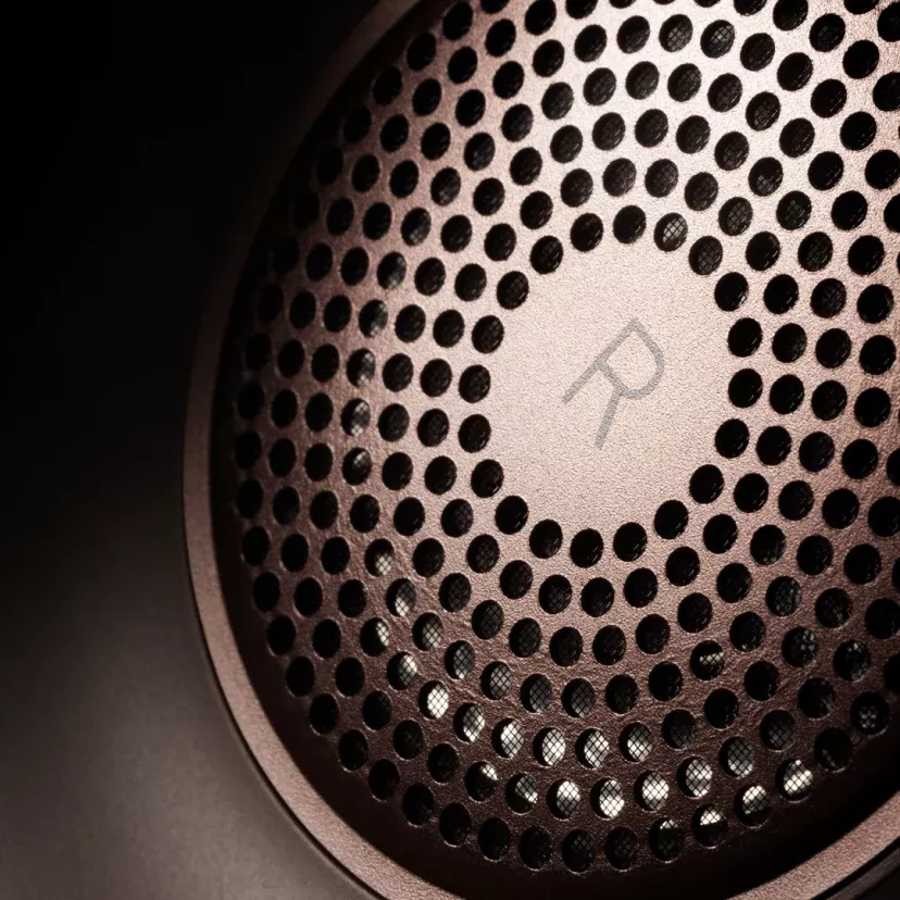 Detail perforované hliníkové mřížky náušníku Beoplay H95 s vyrytým logem Bang & Olufsen