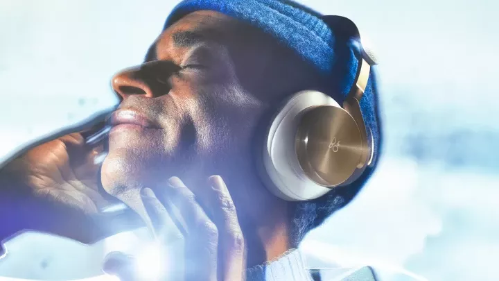 Muž se zavřenýma očima poslouchá hudbu ve sluchátkách Beoplay H95 ve zlatém provedení a dlaní se dotýká náušníku