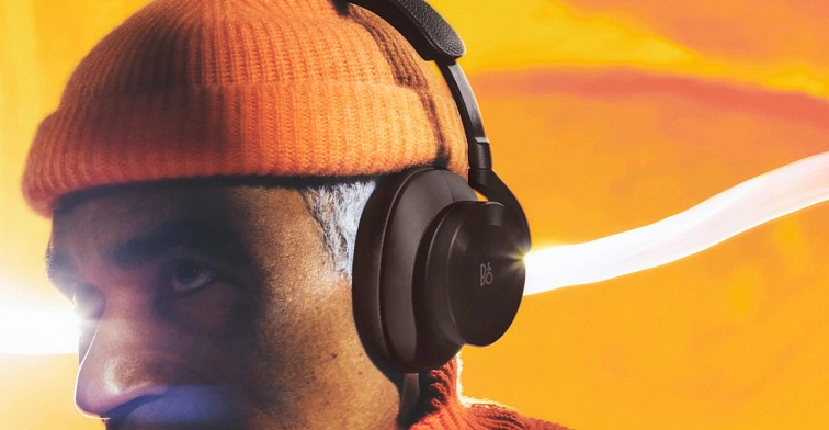Muž v oranžové čepici se sluchátky Beoplay H95 v černém provedení na pozadí oranžového západu slunce
