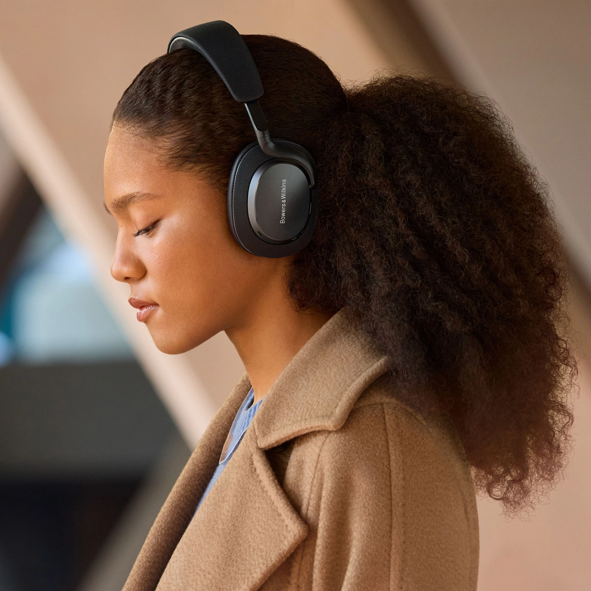 Žena se zavřenýma očima a sluchátky Bowers & Wilkins Px7 S3 v černém provedení, camel kabát