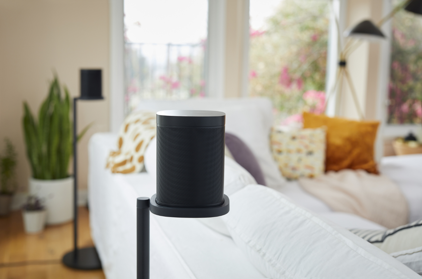 Černý reproduktor Sonos One na podlahovém stojanu vedle bílé pohovky v prosvětleném obývacím pokoji